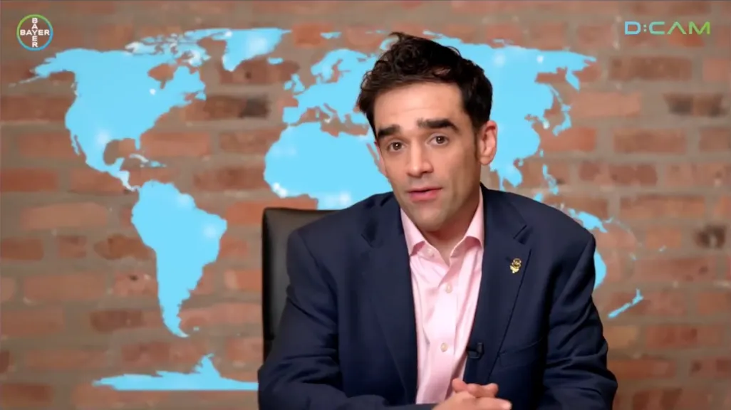 Dr Kishan Rees presenting to camera against a brick wall and a world map, from the D:CAM film.