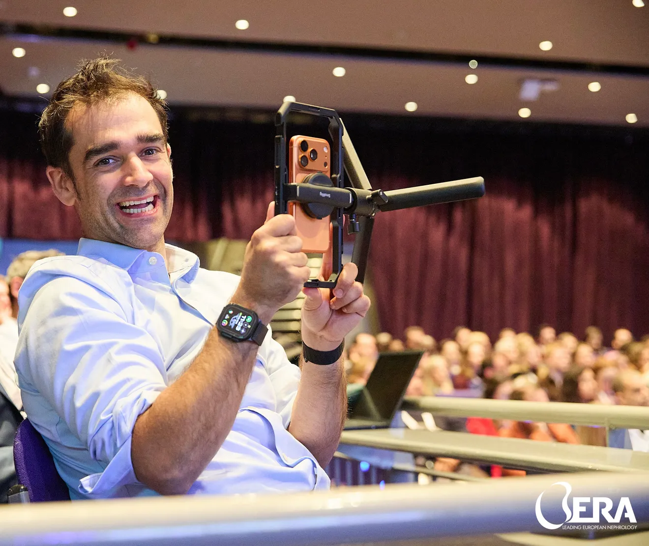 Kishan Rees smiling at the camera while filming a packed conference auditorium with a phone rig.