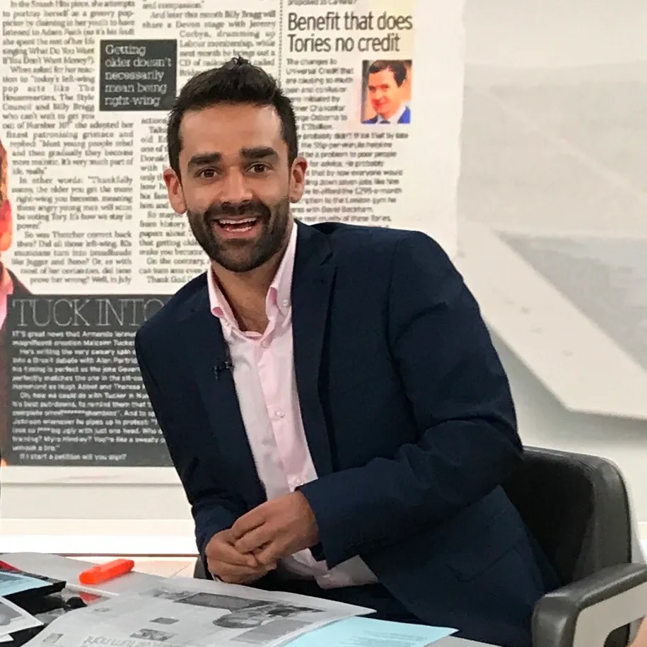 Dr Kishan Rees at a Sky News desk in a navy jacket, smiling to camera in front of a wall of newspaper front pages.